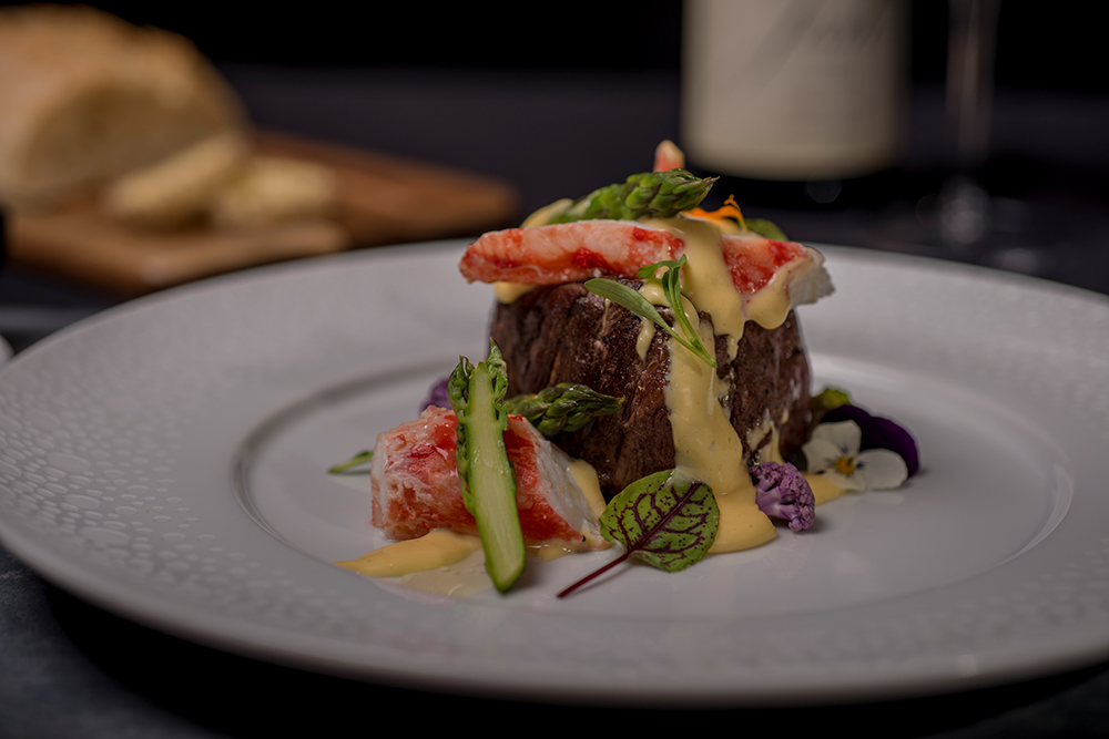 Beef tenderloin on a plate topped with crab meat and creamy sauce, garnished with asparagus, microgreens, and edible flowers.