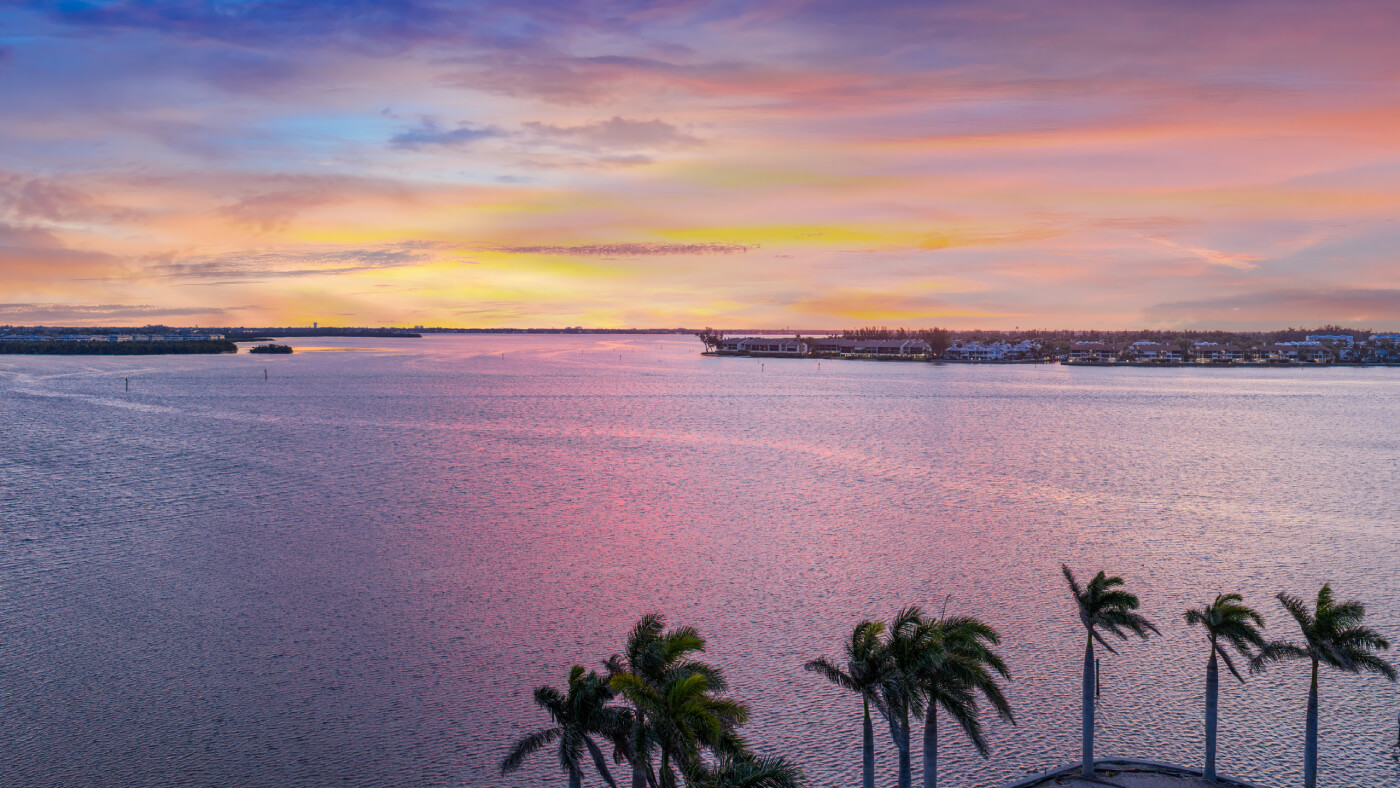 Sunset over calm water with pink and orange sky, palm trees in the foreground and a distant shoreline with buildings.